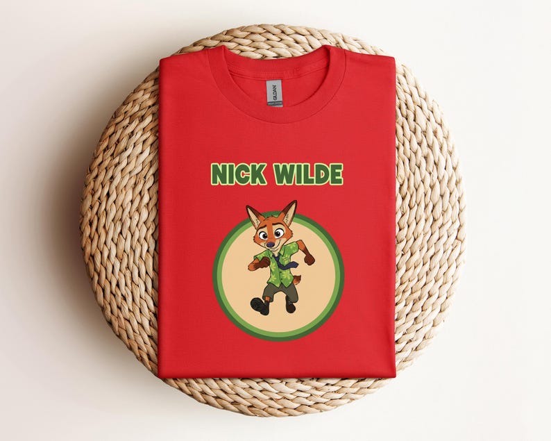 May include: Red t-shirt featuring the text "NICK WILDE" in green above a cartoon fox character in a green and tan circle. The fox is wearing a green shirt and tie. The shirt is displayed on a woven, circular surface.
