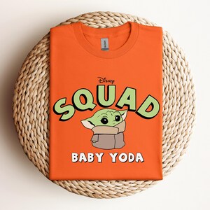 May include: Orange t-shirt featuring the text "Squad" in green with a black outline, the Disney logo, and a graphic of Baby Yoda. The words "Baby Yoda" are printed in white below the graphic. The shirt is folded on a woven mat.