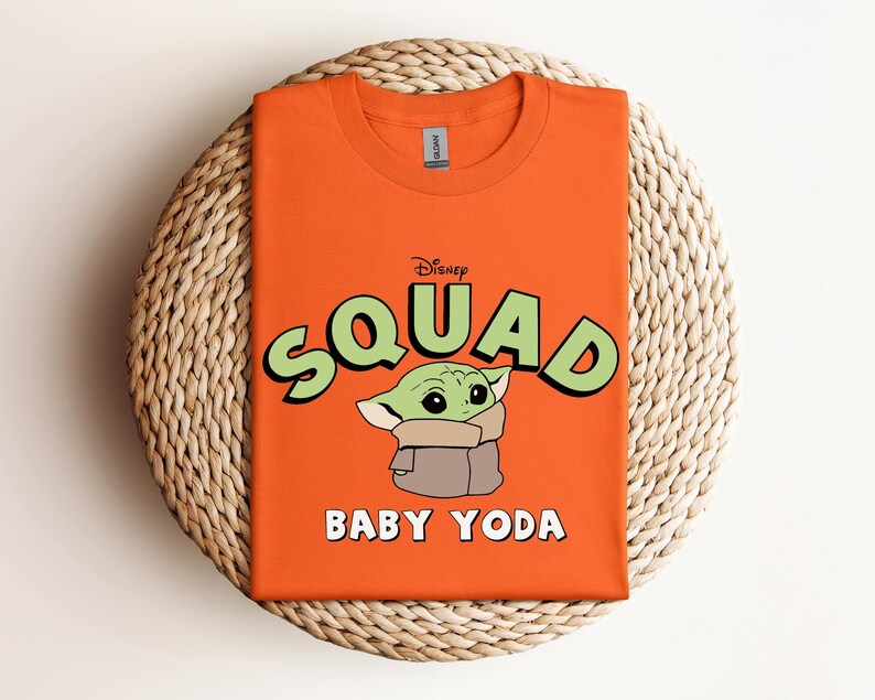 May include: Orange t-shirt featuring the text "Squad" in green with a black outline, the Disney logo, and a graphic of Baby Yoda. The words "Baby Yoda" are printed in white below the graphic. The shirt is folded on a woven mat.