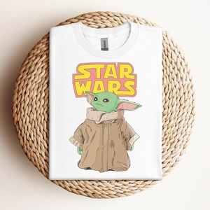 Peut inclure: T-shirt blanc avec un graphisme d'un personnage extraterrestre vert de Star Wars, portant un manteau marron. Les mots "STAR WARS" sont imprim&eacute;s au-dessus du personnage en jaune et rose. Le t-shirt est pr&eacute;sent&eacute; sur un panier en osier.
