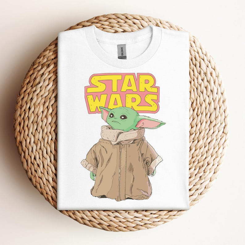 Peut inclure: T-shirt blanc avec un graphisme d'un personnage extraterrestre vert de Star Wars, portant un manteau marron. Les mots "STAR WARS" sont imprim&eacute;s au-dessus du personnage en jaune et rose. Le t-shirt est pr&eacute;sent&eacute; sur un panier en osier.