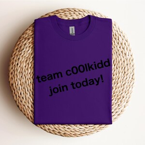 May include: A folded purple t-shirt with the text "team c00lkidd join today!" in black. The shirt is resting on a round, woven wicker placemat.