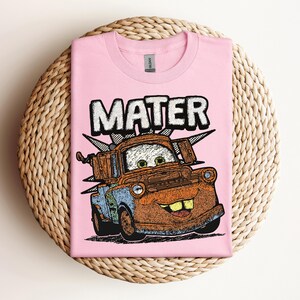 May include: A light pink t-shirt featuring a cartoon image of Mater from the movie Cars. The word "MATER" is printed above the character in a white, outlined font. The graphic is in shades of brown, blue, and orange.