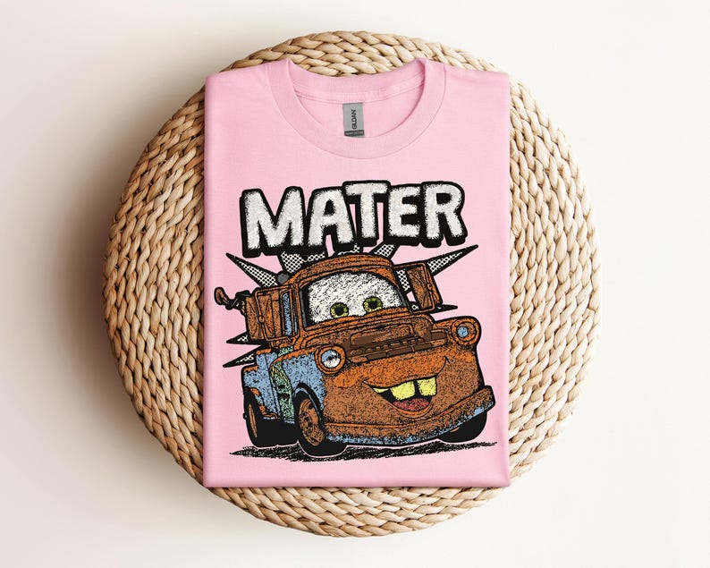 May include: A light pink t-shirt featuring a cartoon image of Mater from the movie Cars. The word "MATER" is printed above the character in a white, outlined font. The graphic is in shades of brown, blue, and orange.