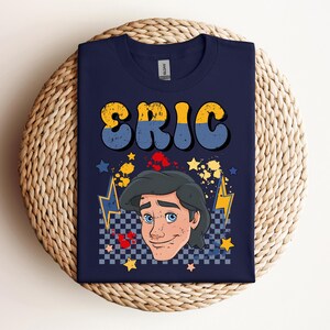May include: Navy blue t-shirt featuring the name "ERIC" in yellow and blue retro font. The shirt includes a cartoon illustration of a character with blue eyes and dark hair, surrounded by stars, lightning bolts, and a checkered pattern.