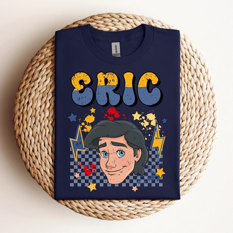 May include: Navy blue t-shirt featuring the name "ERIC" in yellow and blue retro font. The shirt includes a cartoon illustration of a character with blue eyes and dark hair, surrounded by stars, lightning bolts, and a checkered pattern.