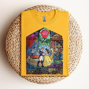 May include: Mustard yellow t-shirt featuring a stained-glass design with a red rose and characters dancing. The graphic includes Belle and the Beast from Beauty and the Beast. The shirt is folded on a woven, circular surface.