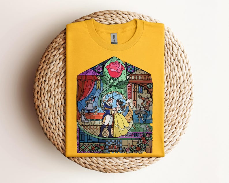 May include: Mustard yellow t-shirt featuring a stained-glass design with a red rose and characters dancing. The graphic includes Belle and the Beast from Beauty and the Beast. The shirt is folded on a woven, circular surface.