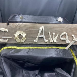 May include: A rectangular wooden sign with the text GO AWAY spelled out using various animal bones. A yellow measuring tape is positioned across the bottom, showing a length of approximately 19 inches from the left edge to the right side.