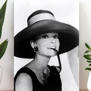 May include: Black and white photograph of a woman wearing a large black hat with a white band. She is wearing a black dress and has a cigarette holder in her mouth.