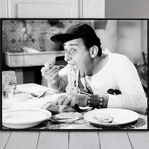 Può includere: Una fotografia in bianco e nero di un uomo con un cappello e una camicia bianca, che mangia spaghetti con una forchetta. È seduto a un tavolo con un piatto di cibo davanti a lui.