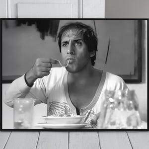May include: Black and white framed print featuring a man eating spaghetti. The man is wearing a white long-sleeved shirt and is seated at a table with a plate of pasta and a glass of water. The background includes an abstract painting.