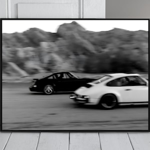 May include: Black and white framed print featuring two classic sports cars racing on a road. The cars are a black coupe and a white coupe, with a blurred mountain range in the background. The image is in black and white.