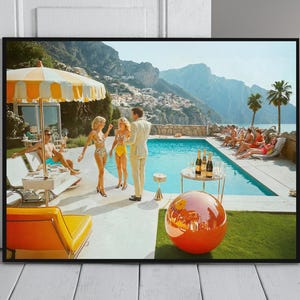 May include: A framed print showing a retro pool party scene. People in swimwear are gathered around a pool with a mountain backdrop. A large orange sphere sits on the grass. A yellow and white striped parasol provides shade.