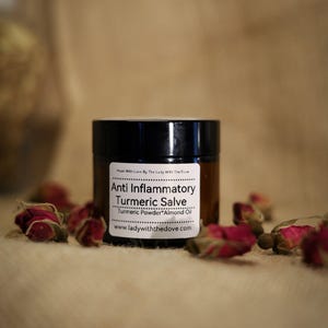 Può includere: Un piccolo barattolo di vetro ambrato con un coperchio nero contenente unguento al curcuma antinfiammatorio. L'etichetta riporta "Made With Love By The Lady With The Dove", "Anti Inflammatory Turmeric Salve", "Turmeric Powder Almond Oil" e "www.ladywiththedove.com".
