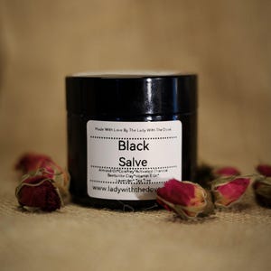 Puede incluir: Un tarro negro con una etiqueta blanca que dice "Black Salve" y enumera los ingredientes, incluyendo aceite de almendras, consuelda, carbón vegetal activado, arcilla bentonita, aceite de vitamina E, lavanda y árbol de té. El tarro está sobre una superficie de arpillera marrón con capullos de rosa secos.