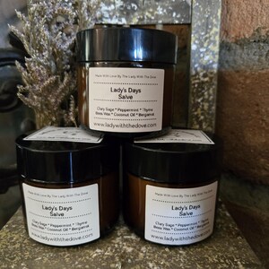 May include: Three brown glass jars with black lids and labels that read "Lady's Days Salve". The labels list the ingredients: Clary Sage, Peppermint, Thyme, Bees Wax, Coconut Oil, and Bergamot. The website address www.ladywiththedove.com is printed on each label.