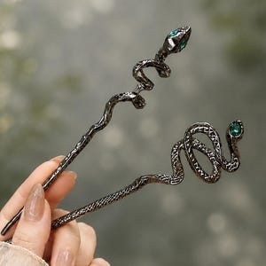 May include: Two dark metallic hair pins featuring a textured, coiled snake design. Each snake head is accented with a small, vibrant green gemstone eye. The pins have a long, slender, and slightly wavy body with a detailed, scale-like surface texture.