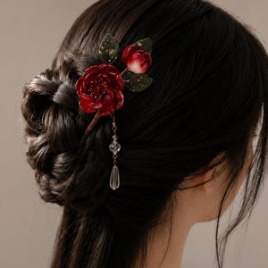 May include: A hair accessory featuring a deep red rose and a smaller bud with dark green leaves. The floral piece is attached to a thin stem and includes a delicate dangling chain with a clear, teardrop-shaped crystal bead at the bottom.