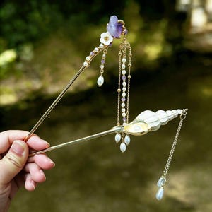 May include: Two ornate hair sticks. One features a purple gemstone, white flowers, and dangling pearls. The other has a white seashell design with dangling pearls. Both are gold-toned and elegant, perfect for special occasions.