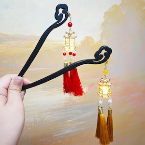 May include: Two black wooden hair sticks with decorative lantern charms. Each charm features a small, lit lantern, red and clear beads, and a tassel. The hair sticks are held against a soft, blurred background.