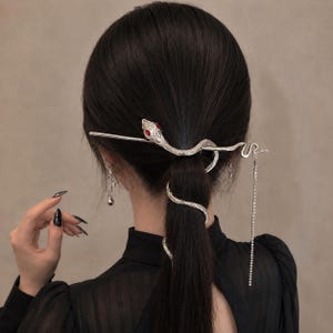 以下が含まれることがあります： 赤い宝石の目を持つヘビの形をした銀色のヘアアクセサリー。ヘビの体がポニーテールに巻き付き、水平に伸びる長いストレートピンと、横に垂れ下がる透明なクリスタルの繊細なチェーンが付いています。