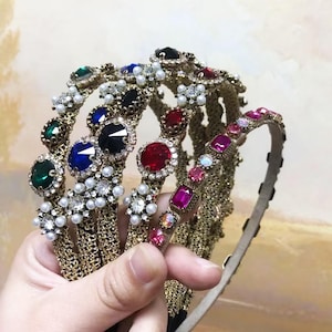 May include: A collection of ornate headbands with jewel and pearl embellishments. The headbands feature a variety of colored gemstones, including emerald green, sapphire blue, ruby red, and black, set in gold-toned metal. Some headbands have chain detailing.