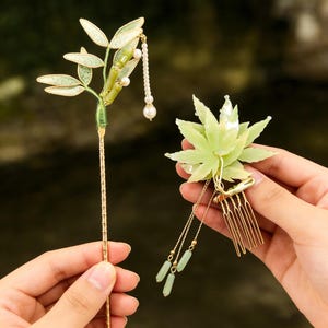 May include: Two decorative hair accessories. One is a gold-toned bamboo-shaped hairpin with green accents and pearl-like beads. The other is a light green flower-shaped hair comb with dangling chains and green beads.