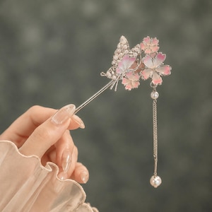 Può includere: Una forcina argentata con un motivo a farfalla e fiori con dettagli in smalto rosa e bianco. Il design è arricchito da piccole perle bianche e include una delicata catenella argentata che termina con una singola perla bianca pendente.