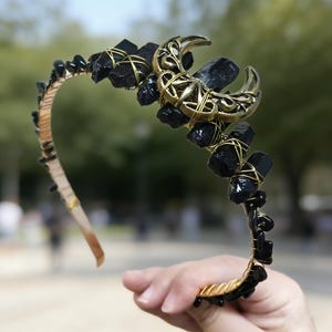 May include: A handcrafted headband featuring a gold-colored crescent moon and black gemstones. The stones are wire-wrapped and arranged along the band, with smaller black beads lining the edges. The headband is designed for special occasions.