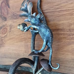 May include: A handcrafted metal door handle featuring a whimsical design. The handle depicts a stylized mouse climbing a branch with leaves and mushroom-shaped accents. The metal has a dark, aged finish, adding to its rustic charm.