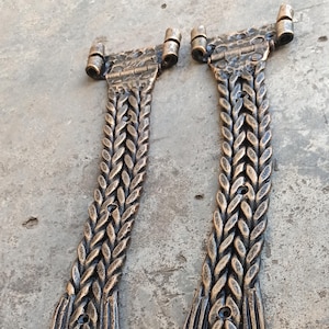 May include: Two bronze-colored, decorative metal hinges. Each hinge features a braided design on the main body, with a rectangular base and a cylindrical top. The hinges are displayed on a gray surface.