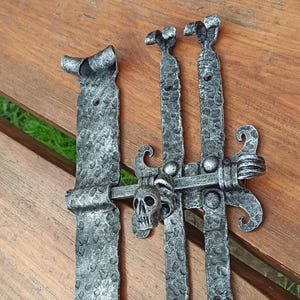 May include: A set of decorative, forged metal gate hardware. The hardware is a dark gray color with a textured surface and a skull design. The set includes hinges and a latch.