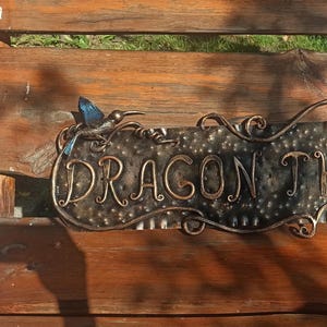 May include: A metal sign with the words "Dragon on the Dirt Inn" in a decorative font. The sign is decorated with metal swirls and two small blue bird figures.