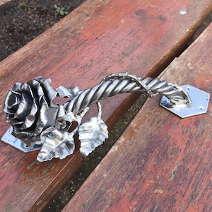 May include: A handcrafted metal door handle featuring a rose design. The handle has a twisted stem and leaf accents, with a silver finish. The rose is attached to a hexagonal base. The handle is resting on a wooden surface.