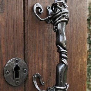 May include: A decorative, dark metal door handle with a unique, twisted design. The handle is mounted on a wooden door with a matching keyhole plate. The design features swirling metal accents, adding a touch of artistry.