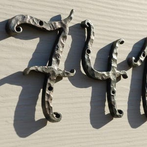 Könnte beinhalten: Ein Set aus vier Hausnummern, die die Ziffern 7, 4, 1 und 1 zeigen. Diese Nummern sind aus dunkelgrauem Metall mit einer gehämmerten, strukturierten Oberfläche gefertigt und haben dekorative, geschwungene Enden. Jede Ziffer hat kleine Montagelöcher.