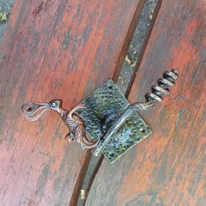 May include: A bronze-colored metal sculpture of a rabbit with long ears, perched on a branch, and a spiraled tail. The rabbit is mounted on a square, textured, green-toned metal plate. The sculpture is set on a wooden surface.