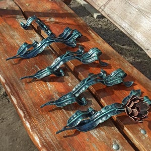 May include: Five decorative metal hooks in the shape of stylized leaves, with a teal green patina finish. The hooks are arranged in a row on a wooden surface. The last hook has a flower design at the end.