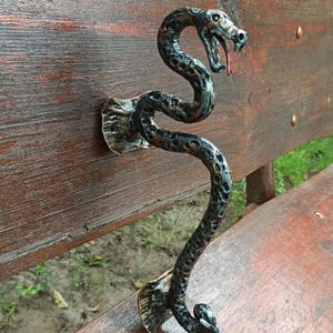 Puede incluir: Un tirador de puerta artesanal con forma de serpiente, de color bronce. El tirador está montado sobre una superficie de madera. La cabeza de la serpiente está con la boca abierta y una lengua roja. El tirador tiene un patrón texturizado en forma de escamas.