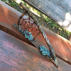 May include: A decorative metal bench ornament with a spiderweb design, a branch-like handle, and leaf accents. The ornament is brown with green and blue accents, resting on a wooden bench.