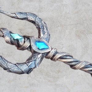 May include: A silver and gold trident with a blue and green gem at the center of the handle. The trident is made to look like it is made of wood and has a twisted handle.