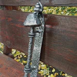 May include: A handcrafted, metal door handle with a mushroom-shaped top and textured finish. The handle is mounted on a textured metal plate and features a decorative, curled design at the top. The handle is made of a silver-colored metal.