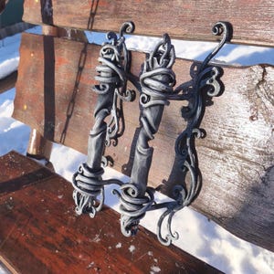 May include: Three ornate, hand-forged metal handles with a dark gray finish. The handles feature intricate scrollwork and twisted metal designs. They are mounted on a weathered wooden surface, suggesting a rustic or vintage aesthetic.