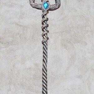 May include: A silver trident with a twisted handle and three prongs. The prongs are decorated with a snake-like design. The handle is adorned with two blue and green gemstones.