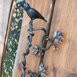 May include: A black metal raven perched on a twisted metal branch with leaves. The raven has blue eyes and is perched on a branch with leaves. The branch is attached to a wall.