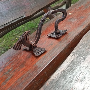 May include: Hand-forged iron gate latch with a bird-like design. The latch features a textured handle and decorative base plates. The metal has a rustic, bronze finish, adding a vintage aesthetic. The latch is designed to be mounted on a wooden surface.