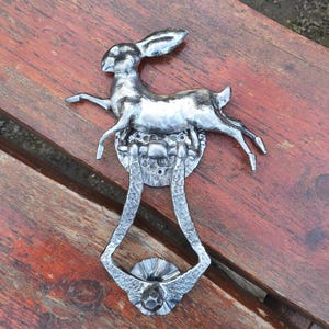 May include: A silver-colored door knocker featuring a leaping hare design. The knocker is made of metal and has a decorative, ornate handle. The hare is the focal point, with its ears perked up and legs extended in mid-leap.