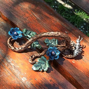 May include: A metal sculpture of a vine with blue flowers and green leaves. A silver-colored ant is crawling on the vine.