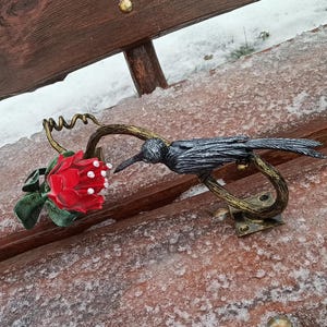 May include: A decorative metal sculpture featuring a bird perched on a curved branch, with a red rose and green leaves. The bird is dark gray, and the rose has white accents. The sculpture is mounted on a wooden surface.